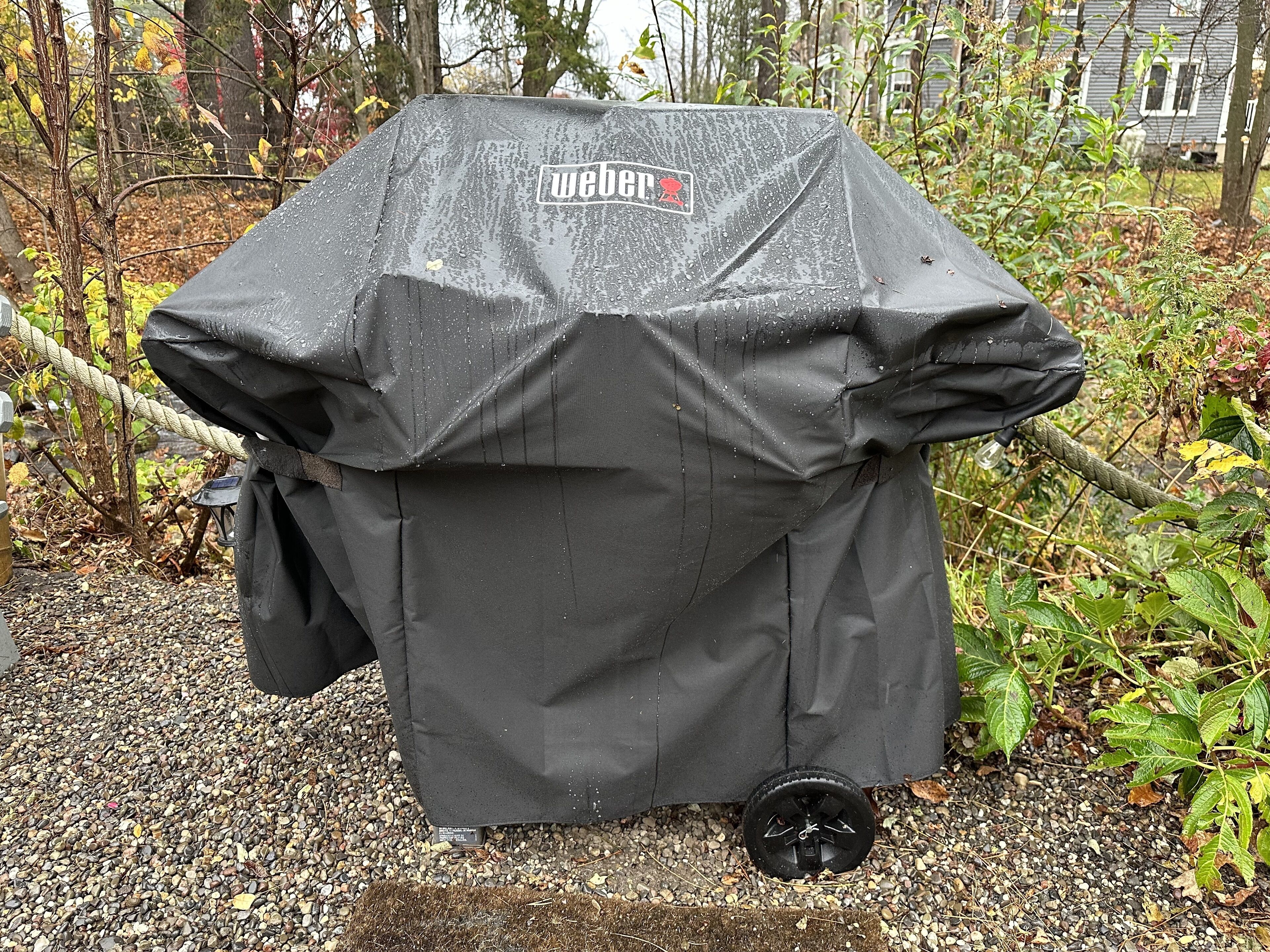 A Weber grill covered in a protective sheet, placed on a gravel surface with surrounding vegetation.