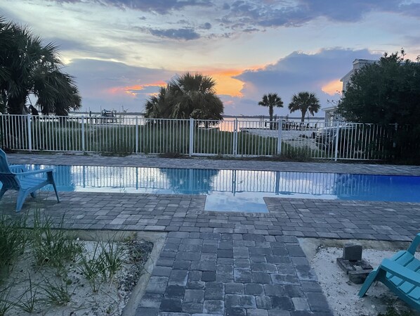 Ein Schwimmbecken mit Blick auf den Sonnenuntergang über dem Meer, umgeben von einem weißen Zaun und Palmen.