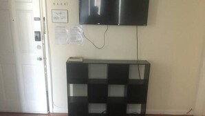 Ein Zimmer mit Holzboden, einem an der Wand befestigten Flachbildfernseher, einer schwarzen Ablage unter dem Fernseher und einem gemusterten Teppich im Vordergrund.