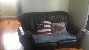 Ein Wohnzimmer mit einem dunkelbraunen Ledersofa, zwei Kissen mit amerikanischer Flagge und Aufdrucken von 'America', einem Holztisch und einem gemusterten Teppich.