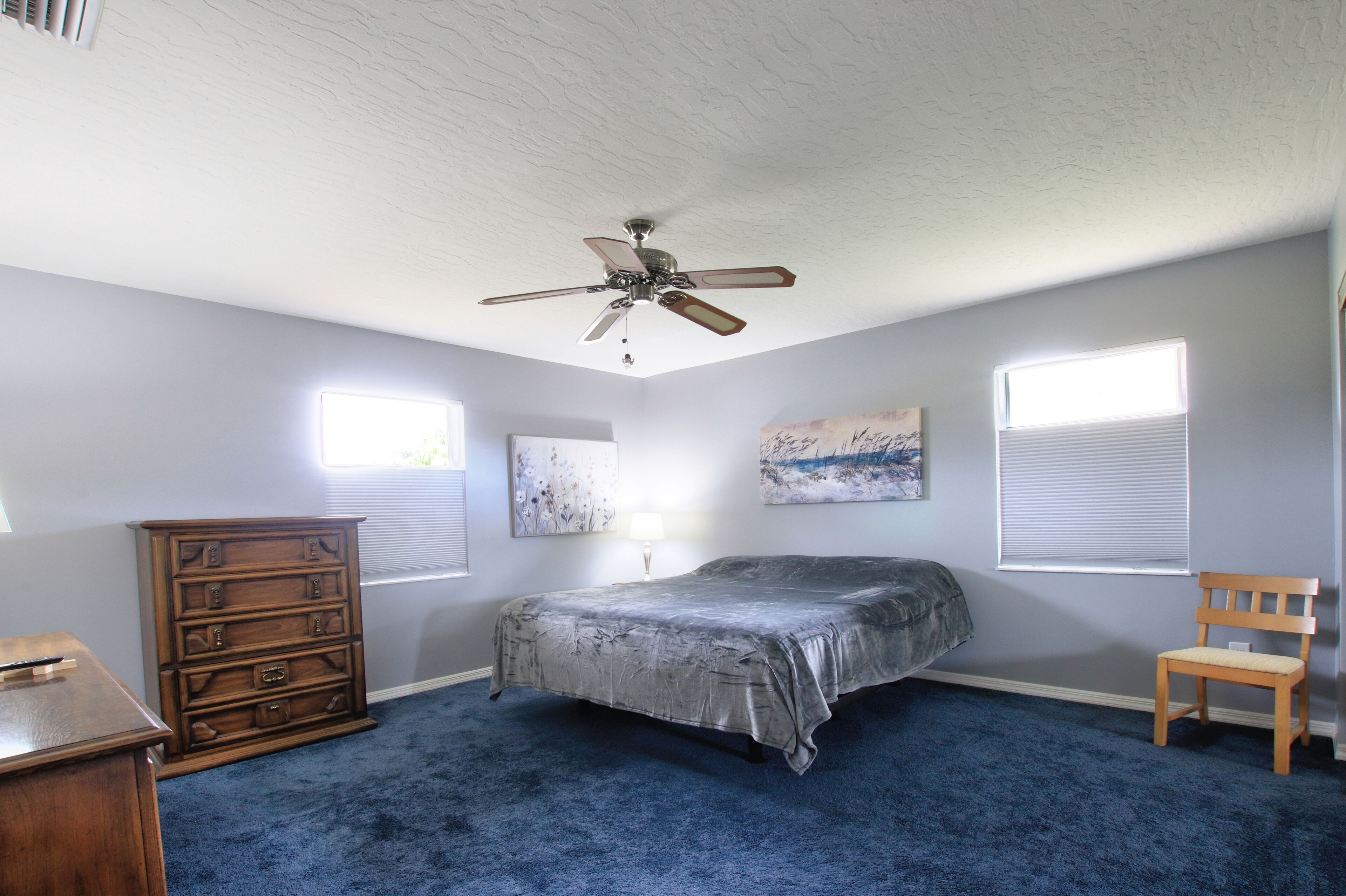 Quarto com cama, cômoda, cadeira, ventilador de teto, obras de arte e janelas.