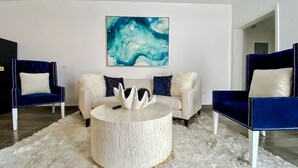 Un salon moderne avec deux fauteuils bleu foncé à revêtement en tissu, un canapé blanc et une table basse ronde dont le plateau est blanc et présente une texture particulière. Un grand tableau abstrait est accroché au mur, au-dessus du canapé.