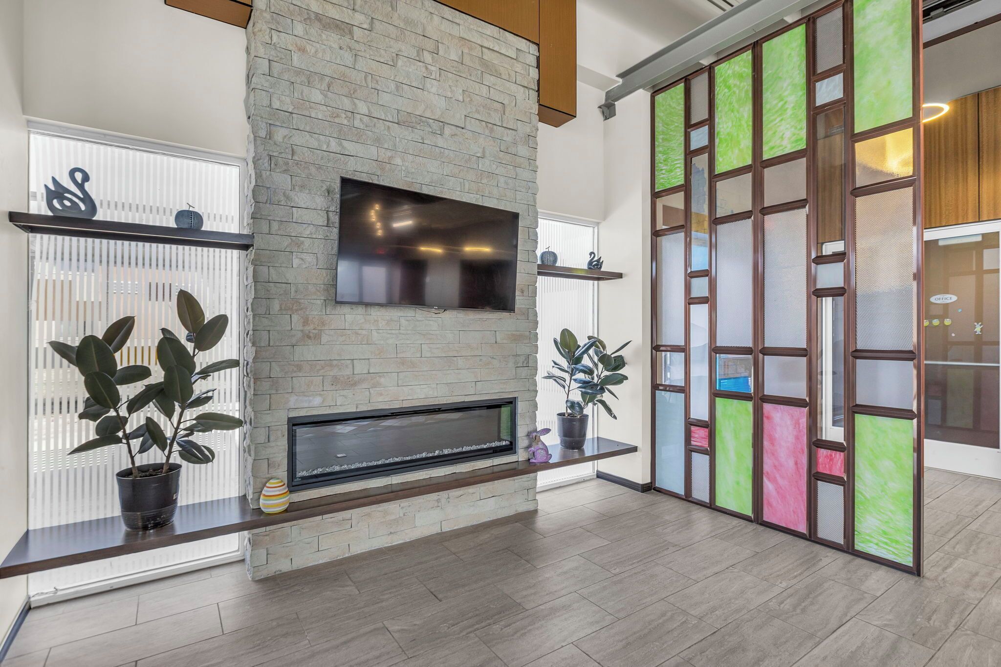 A modern living room with a stone fireplace, a flat-screen TV mounted above it, and a large window with blinds.