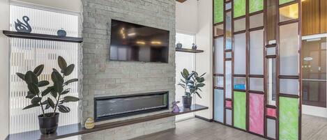 A modern living room with a stone fireplace, a flat-screen TV mounted above it, and a large window with blinds.