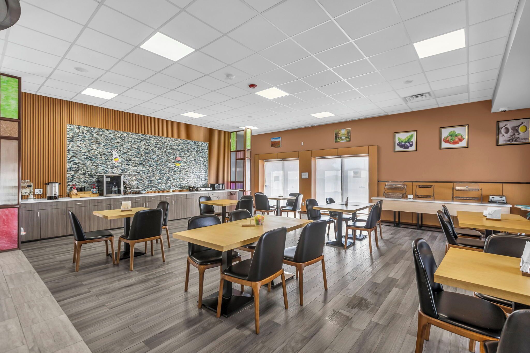 A modern dining area with wooden tables and black chairs, a kitchen area with a stone backsplash, and framed pictures on the walls.