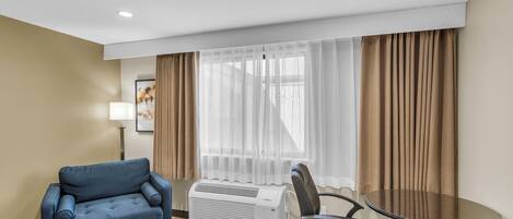 Habitación de hotel con sofá azul, una mesa redonda, una silla de oficina negra, un calefactor portátil y cortinas traslúcidas.