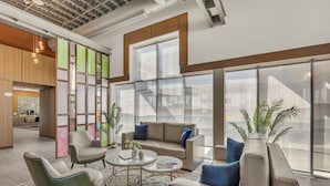 A modern lounge area with a sofa, armchairs, and coffee tables, featuring large windows and indoor plants. - Baymont by Wyndham Kansas City North (North Kansas City)