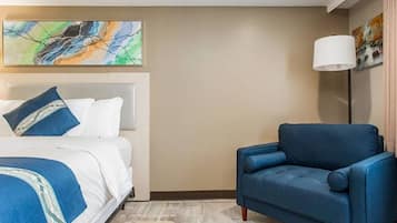 Quarto de hotel com uma cama, uma poltrona azul, um abajur e arte abstrata na parede.