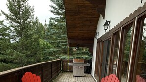A deck with red Adirondack chairs and yellow tables, a grill, and a view of trees.