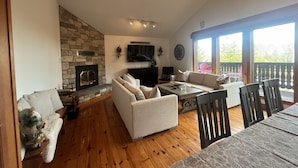 A cozy living room with a stone fireplace, comfortable seating, a flat-screen TV, and large windows offering a view of the outdoors.