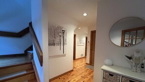 A wooden staircase leading to a hallway with a wooden floor, a white wall with a framed picture, and a round mirror on a white cabinet.