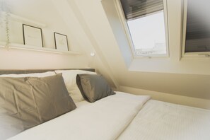 Um quarto com teto inclinado, cama com roupa de cama branca e duas almofadas, janela com persianas e prateleira fixada na parede com quadros.