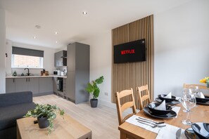 A modern kitchen with grey cabinets, a dining area with a wooden table set for two, and a wall-mounted TV displaying "NETFLIX".