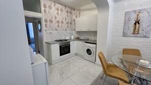 Een kleine keuken met een gasfornuis, oven en wasmachine. Er is een eethoek met een glazen tafel en stoelen.