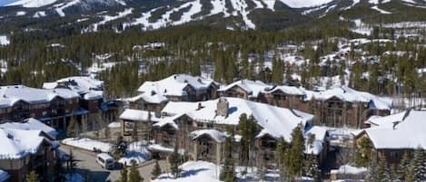 A snow-covered resort with multiple buildings, a ski lift, and a mountainous backdrop.