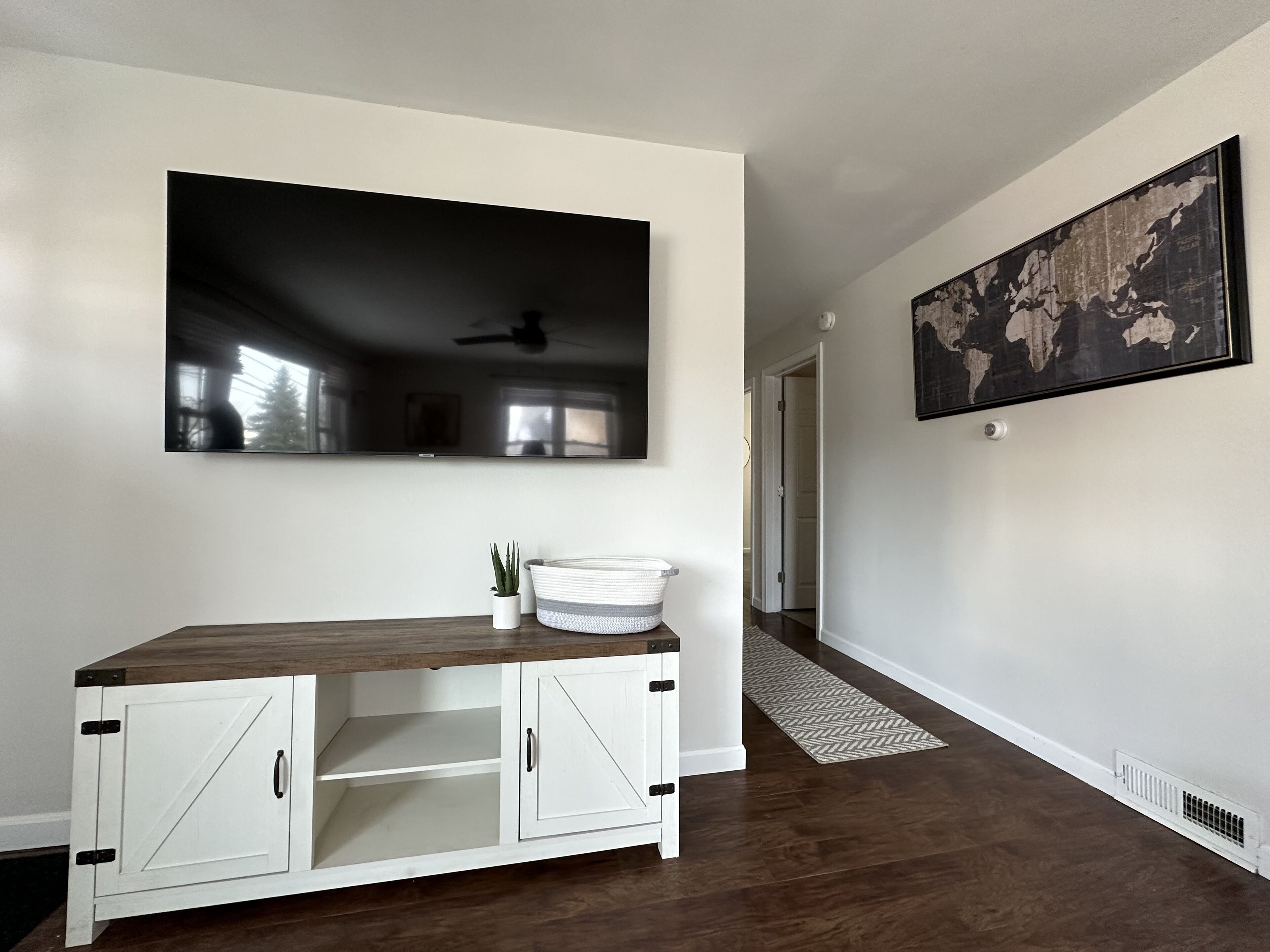 A hallway with a wooden floor, a white cabinet with open doors, a potted plant, a white basket, and a large world map on the wall.