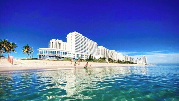 Ett hotell vid stranden med flera höga byggnader, kristallklart blått vatten och en sandstrand.