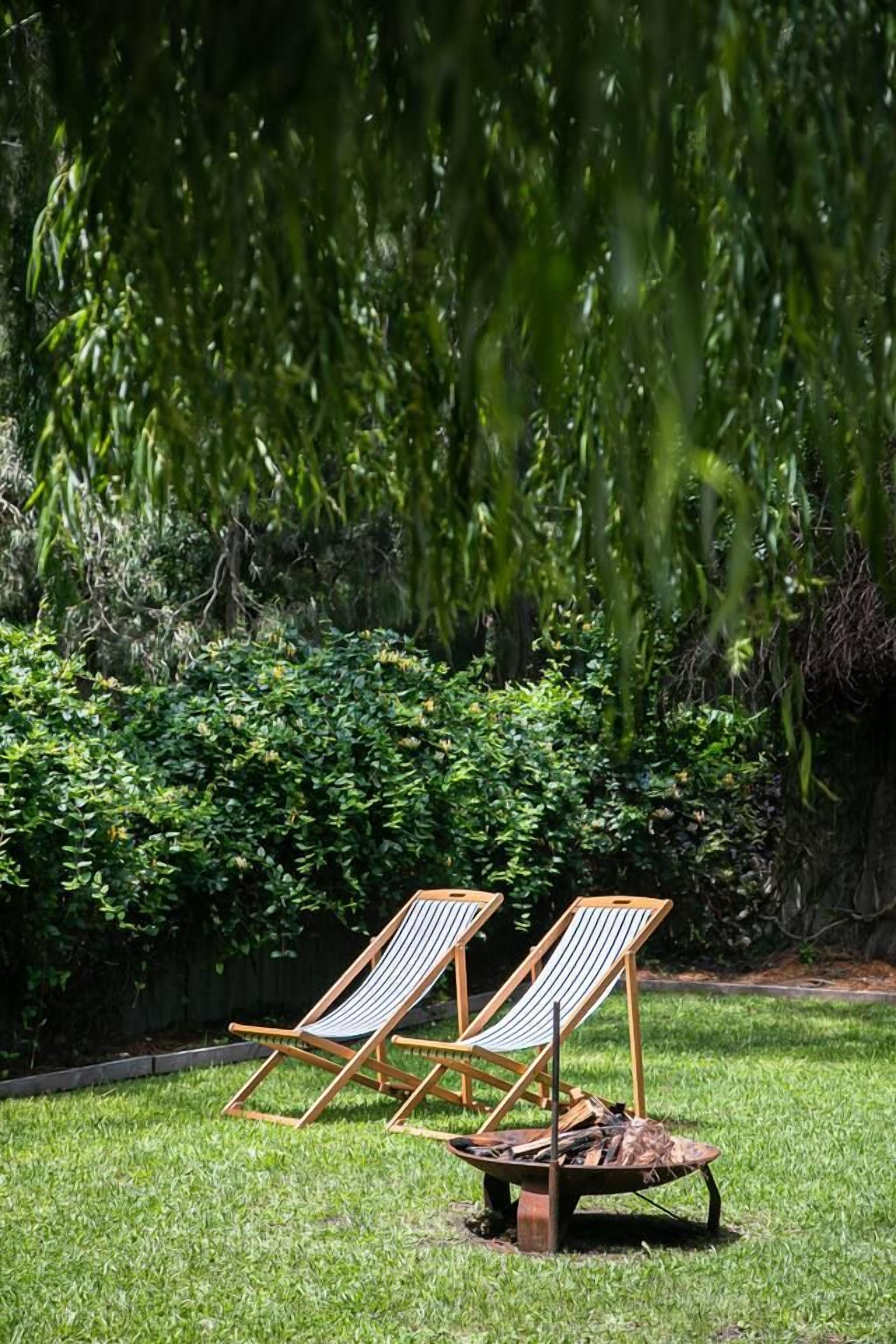 Zwei Holz-Loungesessel mit gestreiften Kissen auf einer grünen Rasenfläche, eine Schubkarre mit Brennholz und dichles Grün im Hintergrund.