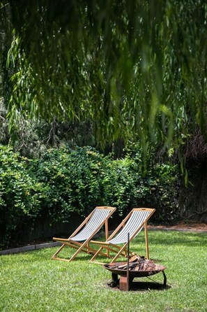Two wooden lounge chairs with striped cushions on a grassy lawn, a wheelbarrow with firewood, and dense green foliage in the background. - Porpoise Palace - Huskisson (Huskisson)