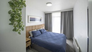 Ein Schlafzimmer mit einem Bett, einem Nachttisch, einer Wandpflanze, einer Deckenleuchte und einem Fenster mit Vorhängen.