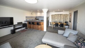 Ein modernes Wohnzimmer mit grauem Sofa, Fernseher und einem kleinen Couchtisch. Es gibt eine Küchenzeile mit Bar und einem Esstisch mit Stühlen.