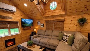 A cozy cabin interior with a fireplace, a sofa, a television, and a window with a view of greenery.