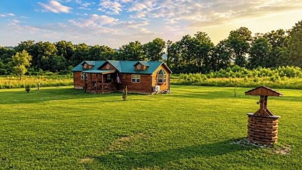 A rustic cabin with a green roof, surrounded by a lush green field and dense trees in the background.
