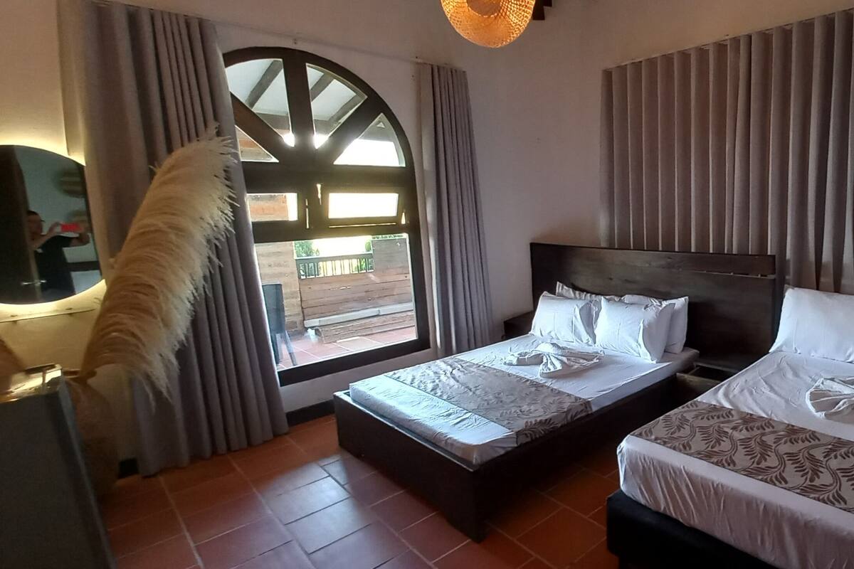 A hotel room with two beds, a large window with curtains, a wooden headboard, and a mirror.