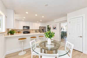 Une cuisine moderne avec des meubles blancs, un îlot central doté de tabourets et une table à manger avec un plateau en verre.