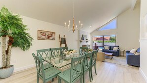 A spacious dining area with a glass table and green chairs, a chandelier, and a large potted plant. - {On IMG Golf Course! WAFFLE BAR/Pool/GameRoom/Beach Gear++ (Bradenton)
