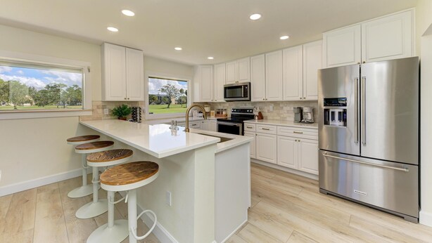 A modern kitchen with white cabinetry, a stainless steel refrigerator, a microwave, and a large island with bar stools. - {On IMG Golf Course! WAFFLE BAR/Pool/GameRoom/Beach Gear++ (Bradenton)