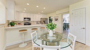 A modern kitchen with white cabinets, a central island with stools, and a dining area with a glass table and chairs. - {On IMG Golf Course! WAFFLE BAR/Pool/GameRoom/Beach Gear++ (Bradenton)