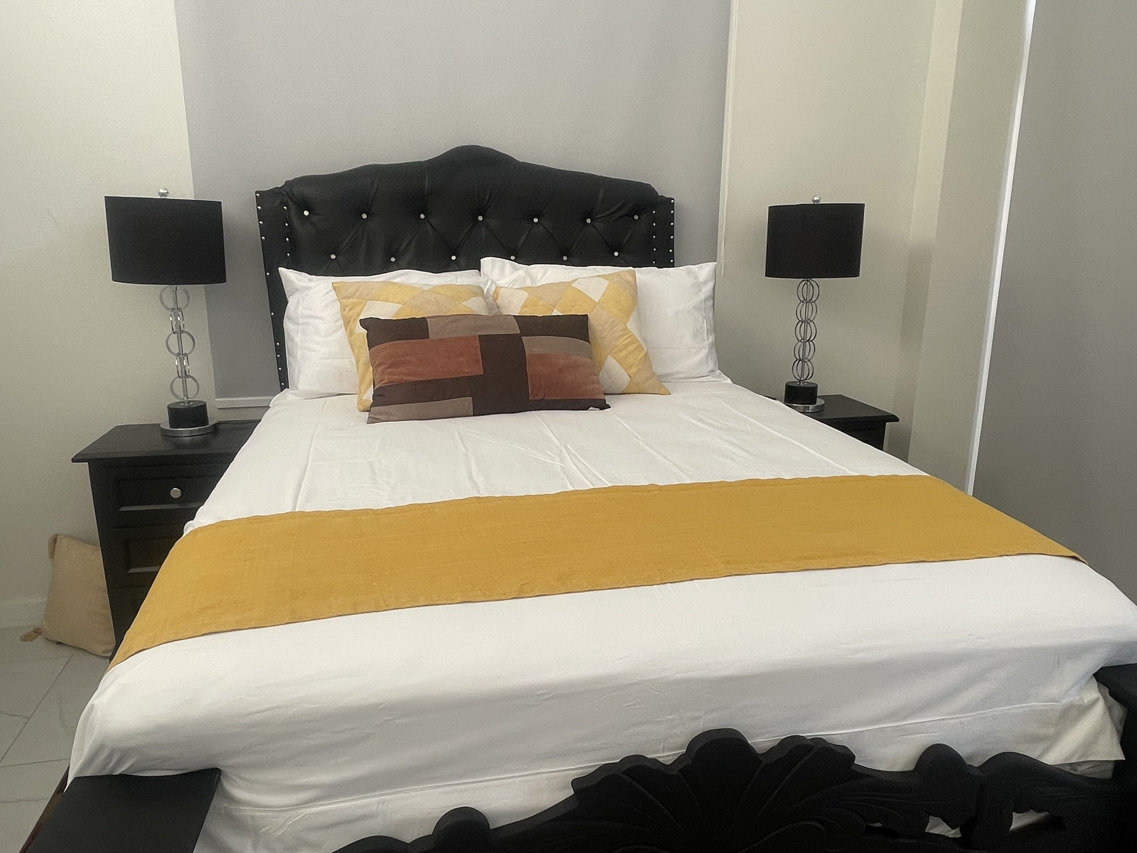 Ein ordentlich bezogenes Bett mit schwarzem Kopfteil, weißer Bettwäsche und einer senfgelben Tagesdecke. Zwei Nachttische mit Lampen und eine kleine Schublade sind vorhanden.