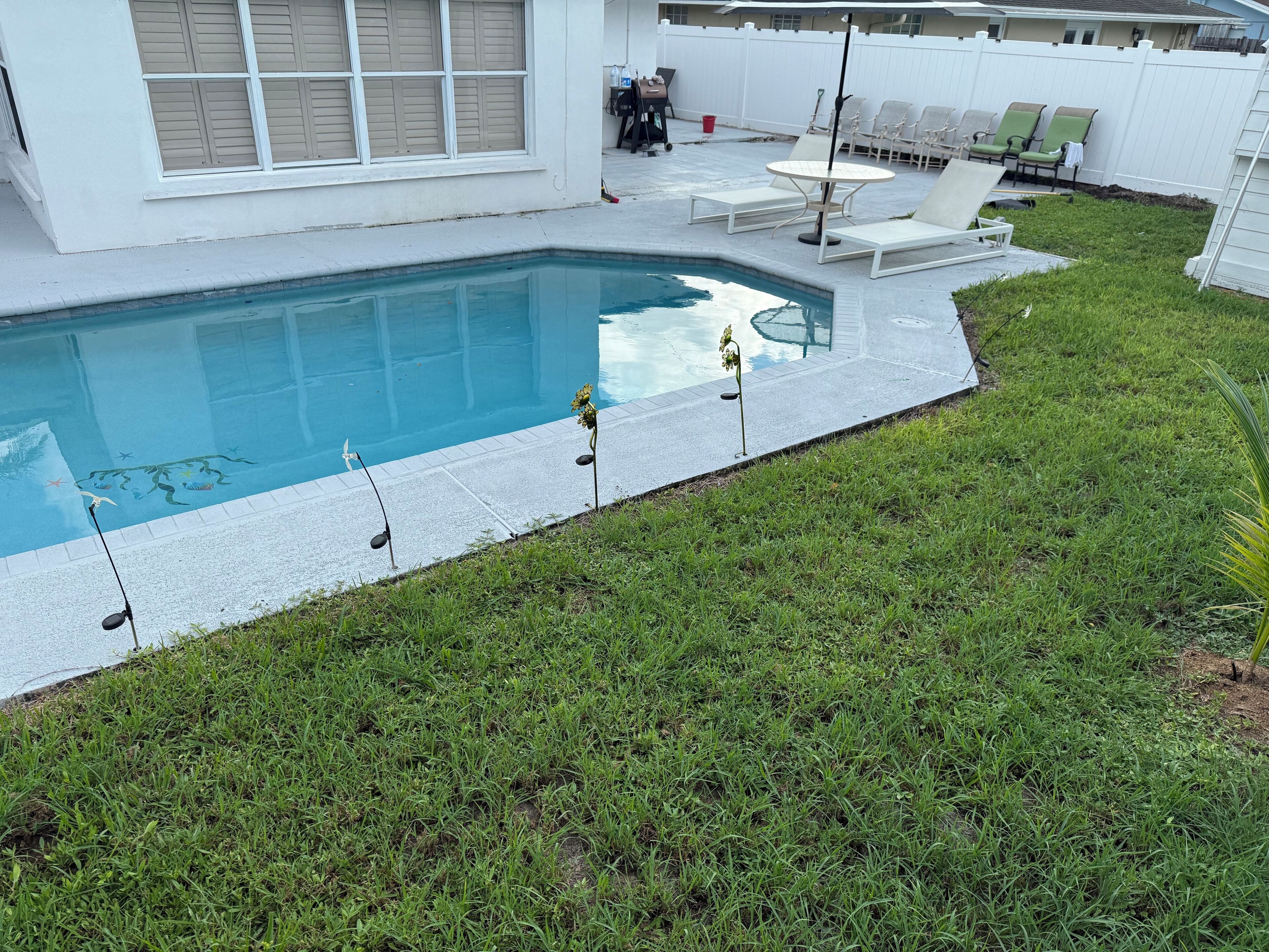 A swimming pool with a tiled deck, surrounded by a grassy lawn and a white fence.