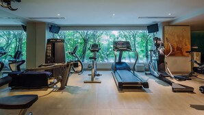 A gym with various cardio machines, a treadmill, and a stationary bike, with a large window offering a view of trees. - Malee Resort Studio 858 (Rawai)