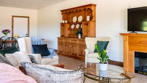 Una sala de estar con un sofá, sillones, una mesa de centro de vidrio, un aparador de madera con platos decorativos y una chimenea.