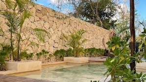 A pool area with a stone wall, surrounded by various plants and a wooden deck. - Your oasis awaits at our luxury penthouse-100% private rooftop with plunge pool, stunning views, 2 additional shared pools on the ground floor and a building elevator just outside the front door.  FREE extras, backup generator and water softener. (Tulum)