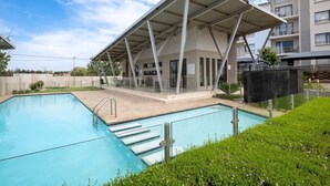 Un edificio moderno con zona de piscina techada, cielo despejado y un complejo residencial al fondo.