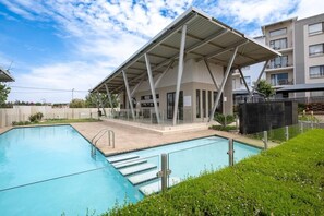 A modern building with a covered swimming pool area, a clear sky, and a residential complex in the background.