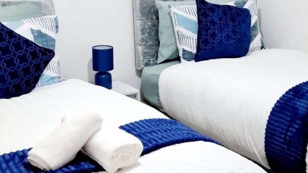 Une chambre d’hôtel avec deux lits, du linge de lit blanc, des touches de couleur bleue et une table de chevet avec une lampe bleue.