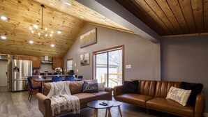 A spacious living area with a high ceiling, wooden beams, a large window, and a kitchen in the background.
