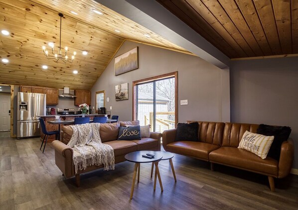 A spacious living area with a high ceiling, wooden beams, a large window, and a kitchen in the background.