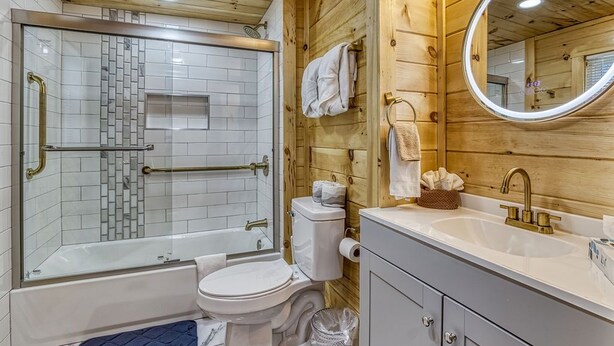 A bathroom with a glass-enclosed shower, a toilet, a sink with a mirror, and wooden walls. - Country Cabin• Fence Area for Dogs• Hot Tub• Games (Kodak)