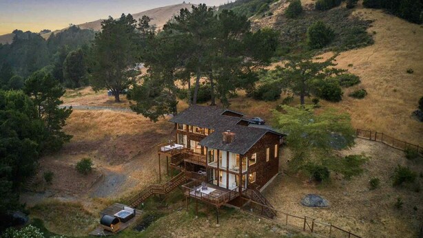 A two-story cabin with a deck, surrounded by a natural landscape with hills and trees. - Sweeping Views, Private Deck,  Outdoor Spa #2 (Nicasio)