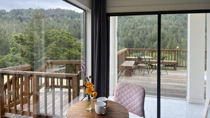 A room with a large window offering a view of a forested hillside. Inside, there's a wooden deck with outdoor furniture, a round table with a vase of flowers, and a cushioned chair. - Sweeping Views, Private Deck,  Outdoor Spa #2 (Nicasio)