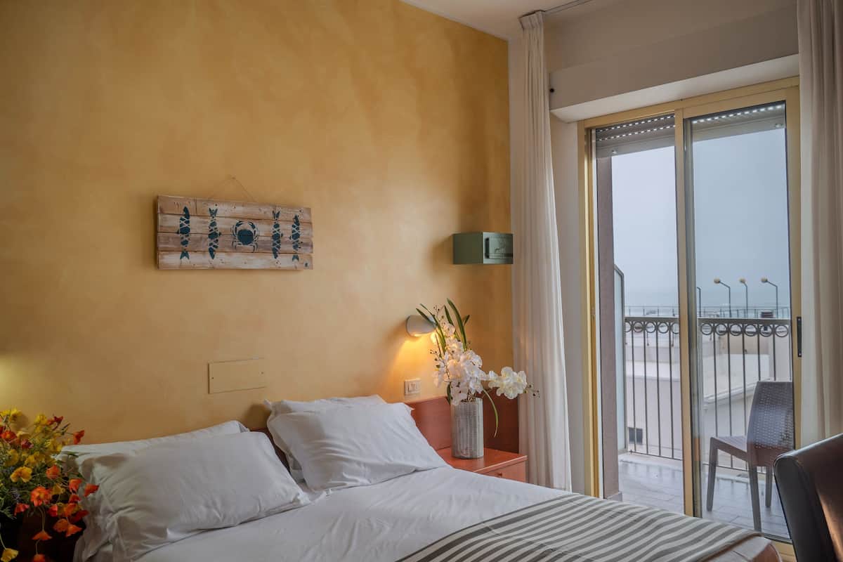 Habitación de hotel con una cama, una mesita de noche con un florero y una obra de arte en la pared, además de vistas al balcón.
