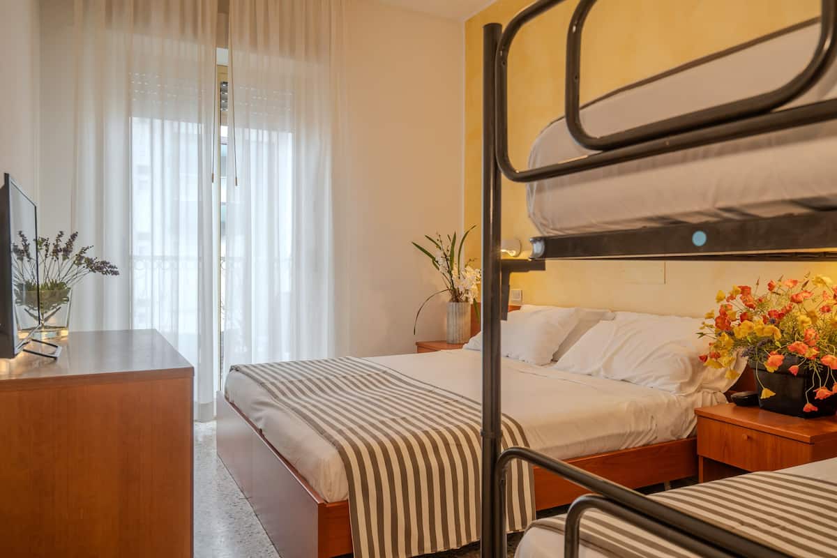 Habitación de hotel con dos camas, un escritorio, un televisor y una ventana con cortinas traslúcidas.