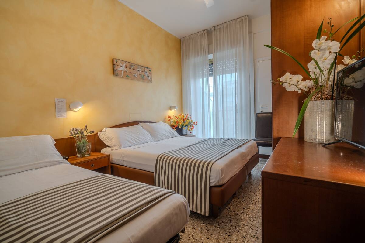 Una habitación de hotel con dos camas, un escritorio de madera, un jarrón con flores y una ventana con cortinas.