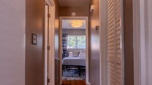 A narrow hallway leading to a bedroom with a bed, a window with curtains, and a ceiling light. - Modern River Retreat Near Airport, Hospitals, Corporations (Wyandotte)