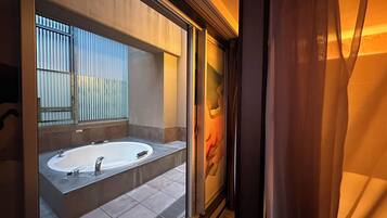 A modern bathroom with a large bathtub, a mirror, and a window with vertical blinds.